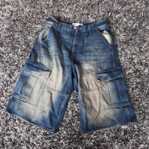 Blå cargoshorts i jeans från New Yorker - Baggy cargoshorts i blått jeansmaterial från New Yorker i storlek S. De har flera stora fickor på sidorna och bak, samt slitna och ljusare partier för en cool, vintage look. Perfekta för dig som gillar streetwear och avslappnad stil. Jag rekommenderar de här till dig som i vanliga fall har storlek XS som jag, själv tyckte jag att den var liten i modellen och en större storlek gav en mer oversize look. 