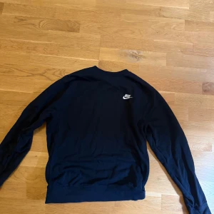 Svart Nike sweatshirt med logga - Svart långärmad sweatshirt från Nike med klassisk vit logga på bröstet. Enkel och stilren design med rund halsringning och mjukt material som passar perfekt till jeans eller träningsbyxor.