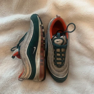 Nike Air Max 97 i grön och silver - Nike Air Max 97 sneakers i grön, silver och grå med reflekterande detaljer och orange insida. Klassisk vågig design, synlig Air Max-sula och snörning framtill. Perfekta för dig som vill sticka ut med en ikonisk och sportig look.