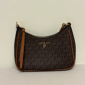 Brun väska Michael Kors - JET SET CHARM CHAIN POUCHETTE, brown/accorn. Använt några gånger. Snygg brun axelväska från Michael Kors med MK-logga över hela väskan. Väskan har guldfärgade detaljer, kedjerem och dragkedja upptill. Tillverkad i syntetmaterial med bruna och beige toner samt läderdetaljer längs kanterna.