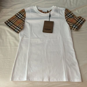 Vit Burberry t-shirt med rutiga ärmar - Snygg vit t-shirt från Burberry med klassiska beige, svarta och röda rutiga ärmar. T-shirten har rund halsringning och är tillverkad i mjuk bomull. Perfekt för dig som vill ha en stilren look med en touch av lyx. Helt ny med tagg