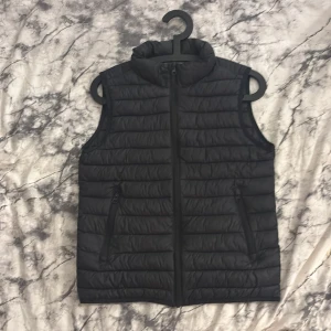 Svart dunväst från McKinley - Svart dunväst från McKinley med quiltad design och hög krage. Västen har två fickor med dragkedja och hel dragkedja framtill. Perfekt för lager-på-lager och outdoor-vibes. Snygg och enkel att matcha med hoodies eller långärmat.