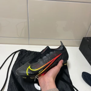 Nike Mercurial fotbollsskor - Säljer ett par grymma Nike Mercurial fotbollsskor i svart med coola röda och gula detaljer. Skorna har en snygg och modern design med snörning för perfekt passform. Perfekta för fotbollsspelare som vill ha både stil och funktion på planen. Använda men i bra skick! Info: Denna sko är utrustad med FG-dobbar och ska enbart användas på naturgräs. Vikt: 205 gram Skick: 8,5/10 Använda: vid 10-20 tillfällen  Inga synliga skador OBS! förutom sulan har ett märke från skohornet.
