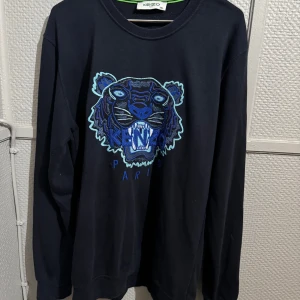 Svart Kenzo sweatshirt med tigerbrodyr - Svart sweatshirt från Kenzo i storlek XXL med ikonisk tigerbrodyr i blå och turkosa toner på bröstet. Tröjan har rund halsringning och långa ärmar. Perfekt statement piece för dig som vill sticka ut med stil.