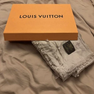 Silvergrå Louis Vuitton halsduk i silke/ull - Lyxig silvergrå halsduk från Louis Vuitton med ikoniskt monogrammönster. Halsduken har fransade kanter och är tillverkad i en blandning av 60% silke och 40% ull, vilket gör den både mjuk och elegant. Perfekt accessoar för att lyfta din outfit.