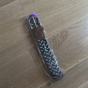 Svart och vit flätad bälte - Säljer ett snyggt flätat bälte i svart och vitt med bruna skinn-detaljer vid spännet. Bältet har ett klassiskt silverfärgat spänne och passar perfekt till jeans eller chinos. Mönstret är randigt och ger en cool touch till outfiten.