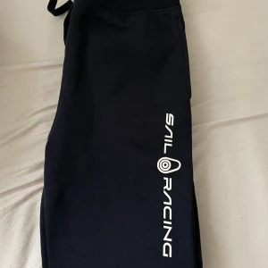 Mörkblå mjukisbyxor Sail Racing - Mörkblå mjukisbyxor från Sail Racing med vit logga längs ena benet. Byxorna har resår och snörning i midjan samt en bakficka. Perfekta för chill eller träning, riktigt sköna och stilrena. Köpt för 699kr förra året!