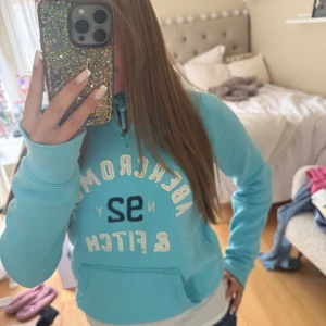 Turkos hoodie Abercrombie & Fitch - Turkos hoodie från Abercrombie & Fitch med broderad vit och marinblå text på bröstet. Klassisk känguruficka, snörning i huvan och mjukt material. Perfekt för chill dagar och snygg till jeans. Det är storlek L men den är väldigt liten i storleken och sitter mer som en S!