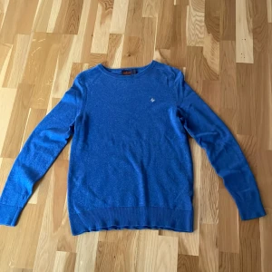 Blå merinoulltröja från Morris - Säljer en blå tröja från Morris i 100% merinoull. Tröjan har rund halsringning, ribbade muddar och en broderad logga framtill. Perfekt för dig som gillar klassisk och stilren look med en modern touch. På tröjan står det att den är i storleken L, men den passar storleken XS.