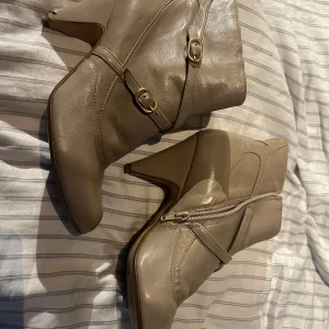 Beige stövletter med klack från Bronx - Snygga beige stövletter från Bronx i skinn med guldfärgade spännen och dragkedja på sidan. Skorna har spetsig tå och hög klack, perfekt för dig som vill ha en stilren och trendig look. Passar till både jeans och kjol. Notera skavanker på undersidan och klacken