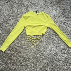Gul croppad topp med snörning H&M - Snygg gul croppad topp från H&M Divided med långa ärmar och trendig snörning i midjan. Toppens material är mjukt och stretchigt, perfekt för att skapa en cool och färgstark look. Passformen är tight och den har en enkel rund halsringning.