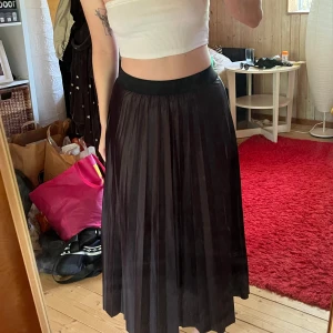 Svart plisserad långkjol - Säljer en svart plisserad långkjol med bred resår i midjan. Kjolen har ett snyggt fall och glansig yta som ger en stilren vibe. Perfekt att styla med crop top eller stickad tröja för en cool look.