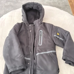 Svart vinterjacka med huva och reflex - Svart vinterjacka från Lyle & Scott med gul broderad logga på ärmen, reflexdetaljer och dragkedja framtill. Jackan har huva, bröstficka med dragkedja och flera smarta fickor. Perfekt för kalla dagar och snygg streetstil. Den har en hål  det kan jag shika mera bilder 
