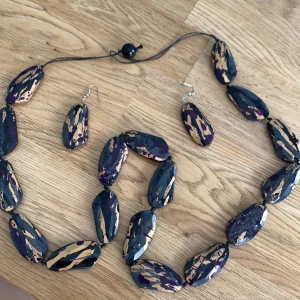Svart och beige halsband & örhängen - Unikt set med halsband och örhängen i stora, ovala pärlor med svart, beige och lila mönster. Pärlorna har en glansig finish och är trädda på svart snöre. Perfekt för dig som vill sticka ut med ett färgstarkt accessoarset.