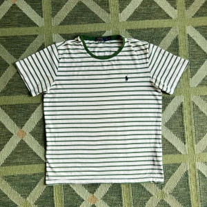 Randig t-shirt från Polo Ralph Lauren - Vit och grön randig t-shirt från Polo Ralph Lauren med klassisk passform. T-shirten har grönt ribbat halslinne och broderad logga på bröstet. Tillverkad i mjuk bomull, perfekt för en chill och stilren look.