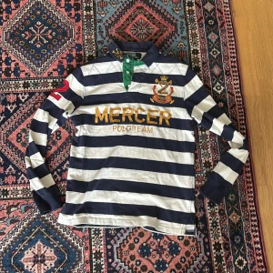 Randig pikétröja Polo Ralph Lauren S - Snygg långärmad pikétröja från Polo Ralph Lauren i marinblått och vitt med breda ränder. Broderad logga och text 'MERCER POLO TEAM' i guld på bröstet, röd siffra på ärmen och grön knappslå. Klassisk krage och skön bomullskvalitet.