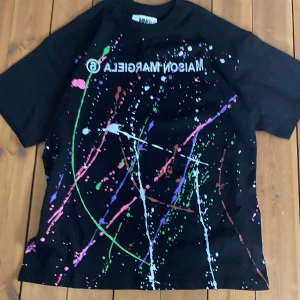 Maison Margiela T-shirt  - Svart t-shirt från Maison Margiela MM6 med coola färgstänk i rosa, grönt, vitt, lila och blått. Tryckt logga på bröstet i vitt. Klassisk passform och rund hals. Perfekt statement-plagg för dig som vill sticka ut.