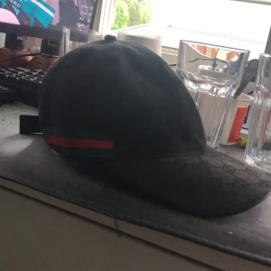 Svart Gucci keps. KAN SÄNKA VID SNABB AFFÄR - Säljer en svart keps från Gucci med klassiskt GG-monogram över hela kepsen. Kepsen har justerbart spänne bak i skinn och en grön-röd rand på sidan. Materialet är bomull och detaljer i skinn, perfekt för dig som gillar exklusiva accessoarer.