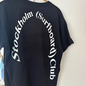 Svart t-shirt Stockholm Surfboard Club - Svart t-shirt från Stockholm Surfboard Club med stor vit text i båge på ryggen. Klassisk passform med rund hals och korta ärmar. Sparsamt använd, nypris runt 1000kr vad jag minns. Mitt pris: 350kr