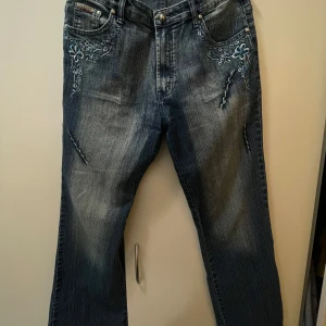 Blå Broderade jeans från Eagame  - Väldigt fina mörkblå jeans från Eagame med ljus tvätt och broderade blommor i blått och vitt på fram och bak. Byxor med fem fickor och raka ben. Bilden med mått blev avkapad vid benen, skriv om du har några frågor!                Längd 104cm, Lår 27cm, Midja 40cm, Höfter 47cm, Ben bredd längst ner 24cm 
