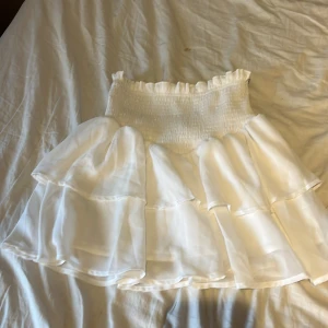 Vit volangkjol med resårmidja - Supersöt vit kortkjol med flera lager volanger och bred smockad resår i midjan. Kjolen är luftig och har en romantisk vibe, perfekt för varma dagar. Materialet känns lätt och flowy, vilket ger en riktigt fin siluett.