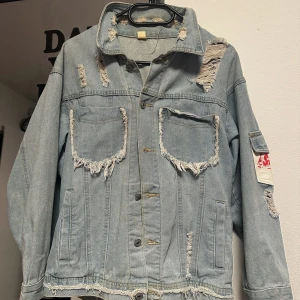 Ljusblå jeansjacka Brazil - Säljer en ljusblå jeansjacka med slitna detaljer och råa kanter. Jackan har två stora bröstfickor, tryckknappar och en stor patch på ryggen med texten 'Brazil' och 'Copacabana' i rött och grönt. Jag har använt flera gånger men det är forfarande ny