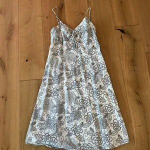 Vit blommig sommarklänning med knyt - Supersöt vit klänning med tunna axelband och svart blommigt mönster. V-ringad med knyt framtill och lätt utställd passform. Perfekt för varma dagar och ger en fräsch och avslappnad vibe.