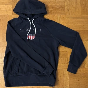 Marinblå hoodie från GANT med tryck - Snygg marinblå hoodie från GANT med broderad logga och sköld på bröstet. Tröjan har huva med vita snören och en stor magficka. Perfekt för dig som gillar klassisk och sportig stil.
