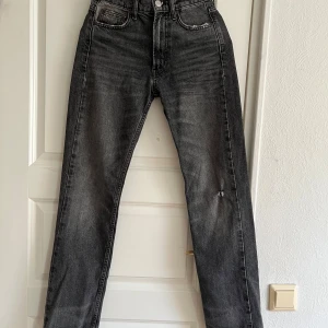 Grå raka jeans från Zara - Snygga grå jeans från Zara i rak modell med hög midja. Jeansen har klassisk femficksdesign, slitna detaljer och knäppning fram. Perfekta till sneakers eller boots för en chill look.