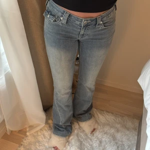 True Religion ljusblå bootcut jeans - Säljer ett par ljusblå jeans från True Religion med bootcut passform. Jeansen har snygga detaljerade bakfickor med lock och silverknappar samt klassisk logopatch bak. Tillverkade i mjuk denim med lätt tvättad look och normal midja. Storlek 27. Inlämnade hos skräddare så kortat ner till min längd 165cm