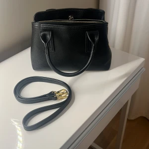 Svart axelväska i skinn med dragkedja - Snygg svart axelväska i äkta skinn med stilrena handtag och avtagbar axelrem. Väskan har guldfärgade detaljer och dragkedja upptill. Perfekt storlek för det viktigaste och passar till många outfits. 27x11x18.