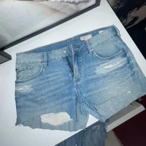 Ljusblå slitna jeansshorts - Snygga ljusblå jeansshorts med råa kanter och slitningar framtill och baktill. Klassisk femficksmodell med knapp och dragkedja. Perfekta för en avslappnad och trendig sommarlook.
