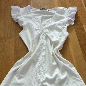 Vit topp med spetsdetaljer - Söt vit topp med v-ringning och spets längs halsringningen och framtill. Volangärmar med broderade spetsdetaljer ger en romantisk känsla. Perfekt för sommarens varma dagar och midsommarfirande.
