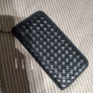 Svart flätad plånbok i skinn - Snygg svart plånbok med flätad design. Plånboken har dragkedja runtom och flera fack för kort, sedlar och mynt. Perfekt storlek för att ha med sig överallt och håller ordning på allt viktigt.
