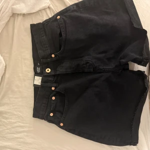 Svarta jeansshorts från 157 DIBA - Svarta jeansshorts från 157 DIBA med råa kanter och klassisk femficksdesign. Shortsen har hög midja, gylf med knapp och dragkedja samt snygga kopparfärgade nitar. Perfekta för en chill sommarlook.