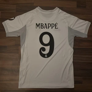 Real Madrid Mbappé matchtröja Adidas S - Säljer en vit Real Madrid 2025/2026 fotbollströja  från Adidas med Mbappé och nummer 9 på ryggen. Tröjan har svarta detaljer, gula linjer, klubbmärke på bröstet och 'Emirates Fly Better' som sponsor. Tillverkad i lätt funktionsmaterial och har korta ärmar.