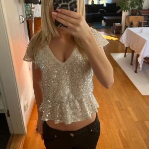 Glittrig topp med volang - Säljer en glittrig topp i silver med v-ringning och volangdetalj i nederkant. Toppen har korta ärmar och är perfekt för festliga tillfällen. Storlek 34. Använd 1 gång
