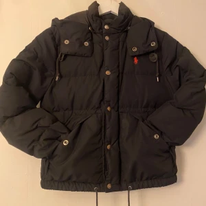 Svart dunjacka från Polo Ralph Lauren - Säljer en svart dunjacka från Polo Ralph Lauren med huva och dragkedja. Jackan har knappar och en broderad logotyp på bröstet. Perfekt för kalla dagar med sin pufferdesign och justerbara detaljer. Det är två stycken små lagningar längst ner på båda armarna.⬛️🟥Hor av dig om du är intresserad🤩passar dig som är 140-150 det går också bra om du är 153.😃