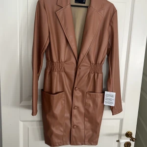 Beige klänning från ASOS ny med prislapp  - Snygg beige kappa från ASOS  i skinnimitation. Klänningen har långa ärmar och knappar framtill. Den har en stilren design med elastiska detaljer i midjan och stora fickor. Perfekt för en trendig look!