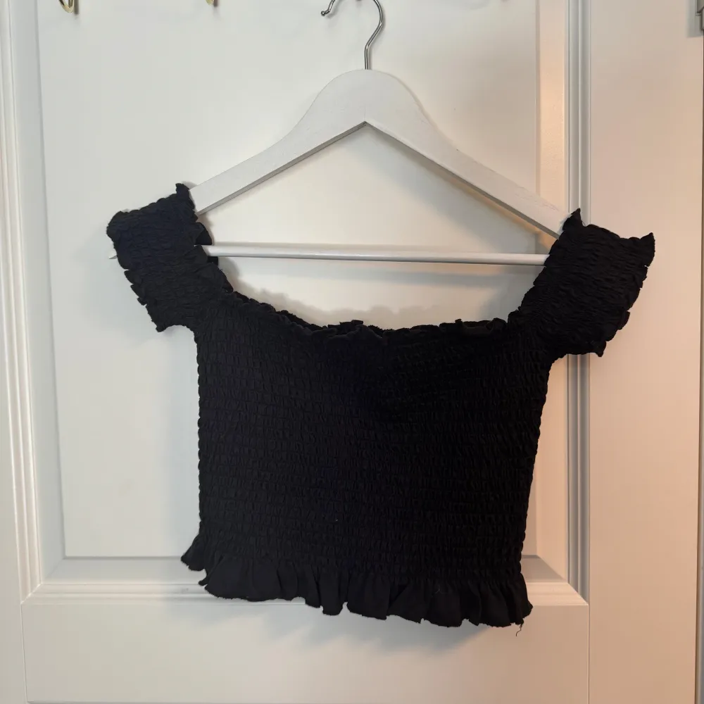 Säljer en svart offshoulder topp från Bik Bok. Toppen är elastisk med en smockad design och har volangdetaljer vid ärmslut och nederkant.. Yläosat.
