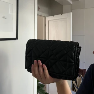 Svart axelväska med kedja - Snygg svart axelväska med quiltat mönster och en elegant guldfärgad kedja som axelrem. 