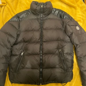 Moncler Chevalier svart - Snygg svart pufferjacka från Moncler med dragkedja och hög krage. Jackan har långa ärmar och en praktisk ficka med knapp på ärmen. Perfekt för kyliga dagar. Mycket fin jacka lite skador finns men inget som syns vid användning 