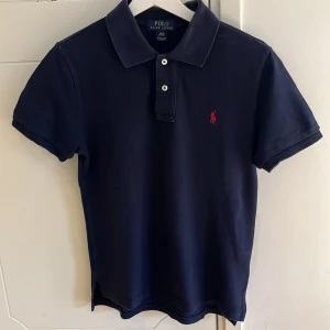Mörkblå pikétröja från Ralph Lauren - Säljer en klassisk mörkblå pikétröja från Ralph Lauren med röd broderad logga på bröstet. Tröjan har korta ärmar och en stilren krage med knappar. Perfekt för en avslappnad men stilfull look. Storlek: L (14-16) Custom fit. Hör av dig för frågor :)