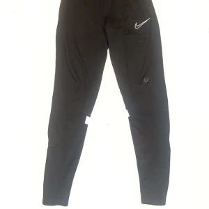 Svarta träningsbyxor från Nike - Säljer ett par svarta träningsbyxor från Nike med en vit logga på framsidan. Byxorna är perfekta för träning. De har även reflekterande detaljer på baksidan av benen. Den har även ett litet bränn märke på knät som gjort att priset sänkts.