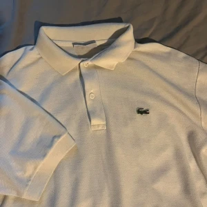 Vit pikétröja från Lacoste - Säljer en klassisk vit pikétröja från Lacoste med den ikoniska krokodilloggan på bröstet. Tröjan har korta ärmar och en knappslå med två knappar. Perfekt för en stilren och avslappnad look.