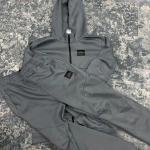 Nike dresss grå - Säljer en stilren grå hoodie med dragkedja och matchande byxor från Nike. Hoodien har en praktisk huva och byxorna har elastisk midja för extra komfort. Perfekt för en avslappnad stil eller träning.