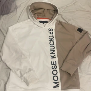Vit och beige hoodie från Moose Knuckles - Säljer en unik hoodie från Moose Knuckles i en cool tvåfärgad design med vit och beige. Hoodien har en stor logotyp på framsidan och en patch på ärmen. Perfekt för en stilren och avslappnad look.