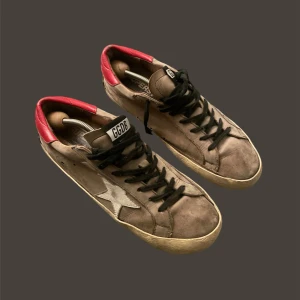 Golden Goose - Snygga sneakers från Golden Goose med en unik design. Skorna har en grå bas med svarta snören och en vit stjärna på sidan. Hälpartiet är i en kontrasterande röd färg.