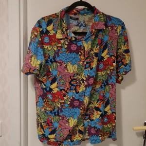 Färgglad kortärmad skjorta från Nazare - Säljer en färgglad och mönstrad kortärmad skjorta från Nazare. Skjortan har ett unikt och livfullt mönster i flera färger som blå, röd, gul och grön. Perfekt för dig som vill sticka ut med en cool och annorlunda stil. Knappar framtill för enkel på- och avtagning.