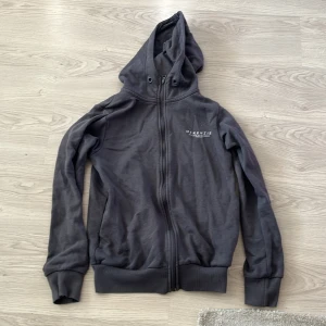 Grå hoodie från McKenzie - Säljer en stilren grå hoodie från McKenzie med dragkedja och justerbar huva. Perfekt för en avslappnad look. Den har långa ärmar och ribbade muddar för extra komfort. Passar bra till både jeans och joggers.